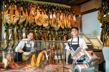 Young woman and man working in jamoneria, standing at counter and cutting ham with special knife.