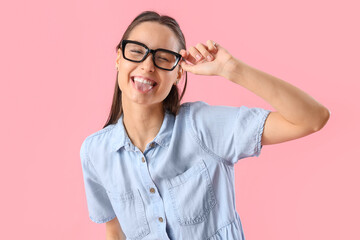 Young woman in eyeglasses showing tongue on pink background