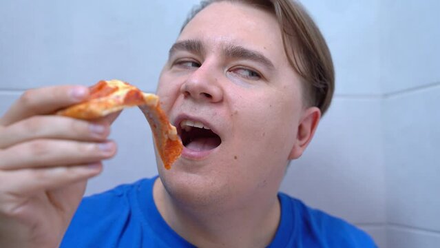 Hand brings slice of thin-crust pepperoni pizza to open mouth, stops, tempts with last piece Man hid in toilet to secretly eat food prohibited on diet, slyly looking sideways, anticipating pleasure