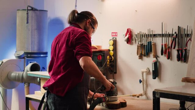 Carpenter collaborating with apprentice, using orbital sander with fine sandpaper to achieve refined finish. Manufacturer and BIPOC colleague using angle grinder on wood block, camera A