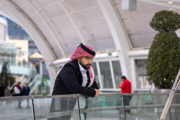 a stylish young man wearing a traditional Arabic headscarf and glasses, leaning against a glass...