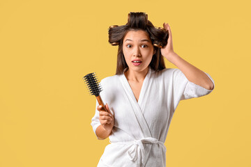 Beautiful young shocked Asian woman in bathrobe with hair curlers and brush on yellow background