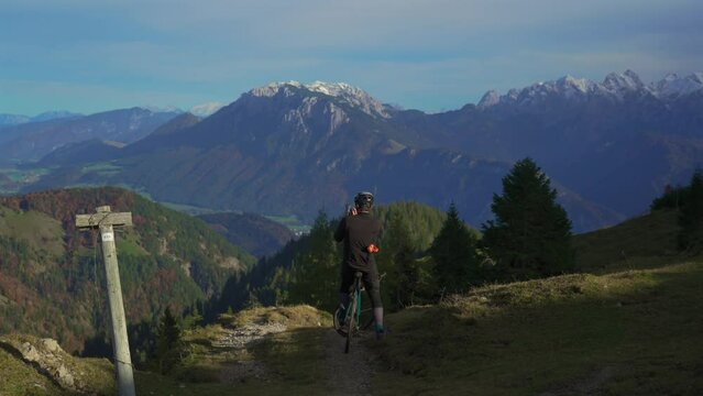 Male cyclist on gravel bike enjoying view and making phono on phone on top of mountain in alps with beautiful view of snowy peaks of mountain range in germany. Sports motivation and inspiration. 