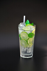 Tall faceted glass of refreshing lemonade with ice and mint on a black background, next to pieces of ripe lime.