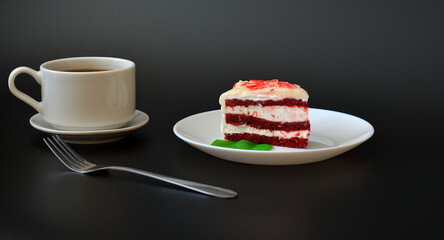 A piece of Red Velvet cake on a plate with mint leaves, a fork and a cup of hot black coffee on a saucer on a black background.