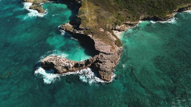 Aerial view of stunning rocky shoreline with waves pounding cliffs under clear, sunny sky, with water so clear it's turquoise. Drone is moving along a circular trajectory.