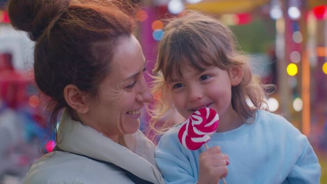 Mom holds and kisses her little daughter who is eating a big candy in an amusement park