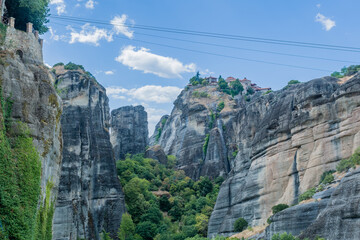 Ancient monastery perched on top of a high cliff surrounded by lush greenery under a blue sky with clouds in Meteora, Greece
