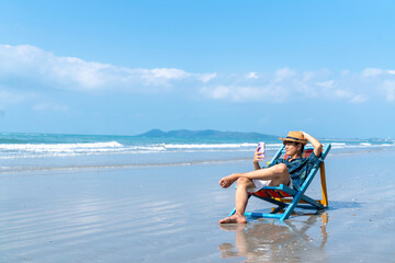 Happy Asian man enjoy outdoor lifestyle travel ocean on summer holiday vacation. Handsome guy relaxing and using smartphone for online shopping or chatting at tropical island beach in sunny day. - obrazy, fototapety, plakaty