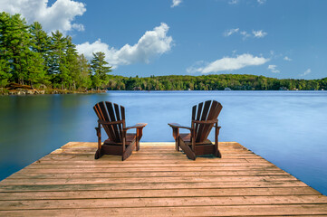 Two Adirondack chairs sit on a wooden dock beside a calm lake. Rustic cottages nestled among lush...