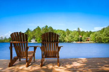 Two Adirondack chairs sit on a wooden dock, overlooking a tranquil lake. On the opposite shore, a...