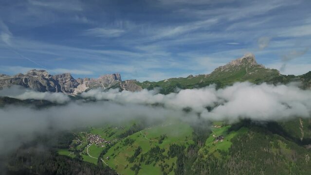 Drone flies forwards slowly turning right, revealing Sass de Putia in cloud-kissed Dolomites. Chalets dot lush hills as drone drifts above them, capturing serene village. La Val, Italy. LuPa Creative.