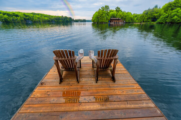 Two Adirondack chairs with a mug of coffee are placed on a wet wooden pier overlooking a tranquil lake in Muskoka, Canada. It's early morning at the cottage, with a vibrant rainbow adorning the sky.
