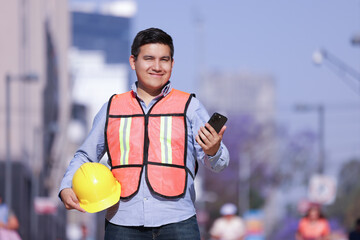 mexican working man smiling holding helmet and cell phone with city background