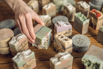 A lot of pieces of fragrant, beautiful natural soap on a wooden table in the workshop and the hand...