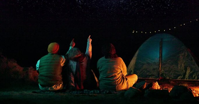 Friends, tent and camping with stars at night for adventure, holiday fun and outdoor activity. Campsite, people and pointing to sky with rear view, leisure in nature and travel with bonfire in woods