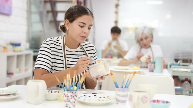 Skilled interested young female artist sitting at table in pottery workshop, painting designs on ceramic cup. Art craft activity concept