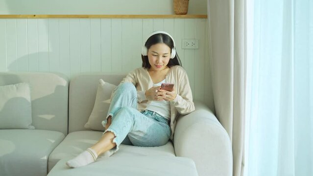 Happy young Asian woman with mobile phone listening music in headphones and relaxing on sofa at home