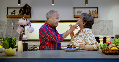 Indian Asian Hindu older lover gen x couple pair sitting modern kitchen pastry cake cream put apply...