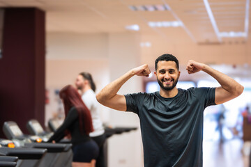 Joyful man showing off muscles with a flex at the gym, companions working out behind. 