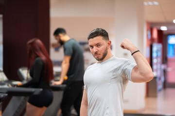 Muscular man in white tee flexes his bicep, with focused gym-goers behind him. 
