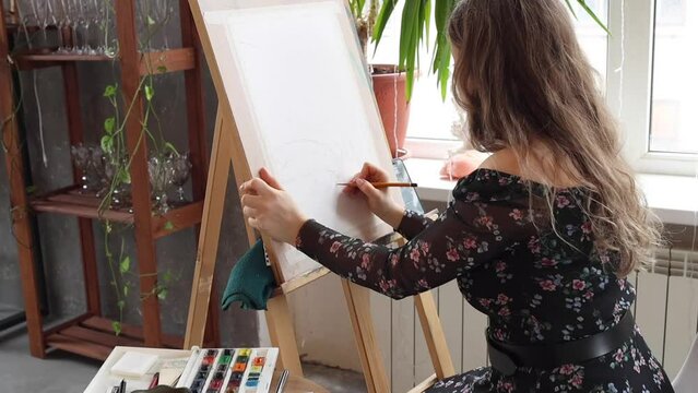 Beautiful woman artist painting with pencil on paper and wooden easel. Female working in art studio in process. Blonde girl with long hair wearing black floral dress