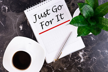 Just Go For It motivational message is written in a notebook lying on a grey table with a pen and a...