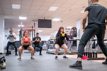 A diverse group of gym-goers engages in a kettlebell squat session with personalized coaching from a fitness instructor in a well-equipped gym. 