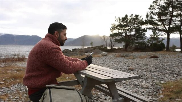 A man in a cozy fleece jacket enjoys a moment of solitude at a picnic table amidst a tranquil pebbly landscape.