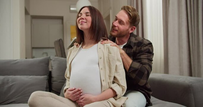 Happy man with brown hair with stubble in a plaid shirt doing a shoulder massage for his pregnant brunette wife while sitting on a gray sofa in a modern apartment