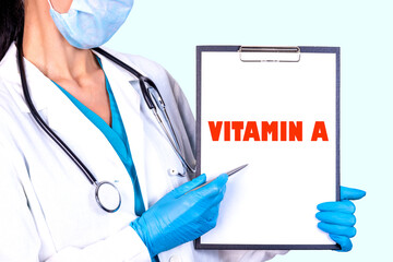 A doctor in blue gloves holds a tablet with a clip labeled VITAMIN A and shows the pen on the...