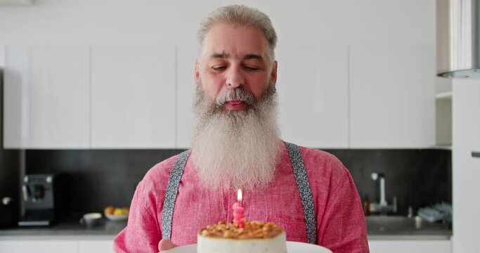 Portrait of a happy elderly man with gray hair with a lush beard in a pink shirt who holds a cake with a burning candle in front of him and blows it out in honor of his birthday in a modern kitchen