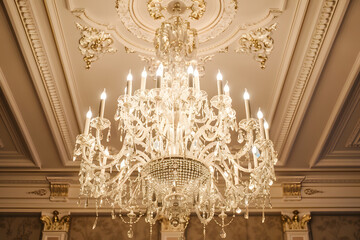 A grand crystal chandelier hangs majestically in a luxurious room, its lights casting a warm glow that highlights the ornate ceiling details..