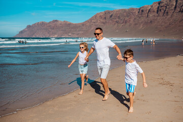 Father with two kids running on beach. Young family enjoying sandy beach, looking at crystalline sea in Canary Islands. Concept of beach summer vacation with kids.