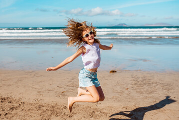 Jumping girl on beach. Smilling blonde girl enjoying sandy beach, looking at crystalline sea in Canary Islands. Concept of beach summer vacation with kids. - obrazy, fototapety, plakaty