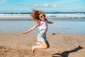 Foto op Plexiglas Jumping girl on beach. Smilling blonde girl enjoying sandy beach, looking at crystalline sea in Canary Islands. Concept of beach summer vacation with kids. © Halfpoint