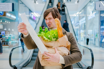A man in a casual outfit is visibly perplexed while looking at a lengthy grocery receipt, holding a...