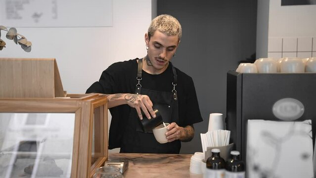 Handsome barista guy work coffee shop. Tattooed hipster man. Male face smile. Joy young adult person cafe house. Stylish dyed hair waiter make hot tasty drink order. Latte art cafeteria. Bar staff job