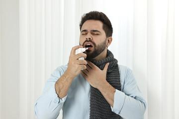 Young man with scarf using throat spray indoors