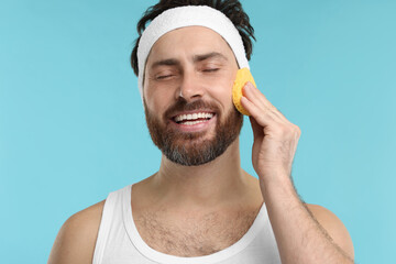 Man with headband washing his face using sponge on light blue background