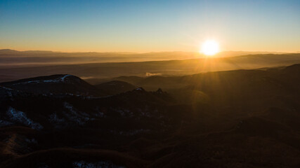 Naklejka na ściany i meble The sun rises over a serene mountain landscape, casting a warm glow over the misty valleys and undulating hills, captured in an expansive aerial view.