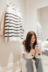 Korean teenage girl takes a selfie in new clothes in the mirror and sends it to her friend via phone in a shopping mall. Retail and consumerism. Sale promotion and shopping concept. Part of a series - 779467143