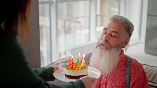 A happy man with gray hair and a lush beard in a pink shirt blows out four candles and on a small cake during his birthday and congratulations from his adult daughter in a modern apartment
