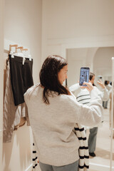 Korean teenage girl takes a selfie in new clothes in the mirror and sends it to her friend via phone in a shopping mall. Retail and consumerism. Sale promotion and shopping concept. Part of a series - 779468352