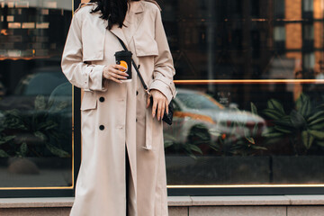 Street style photo, cropped young woman wearing beige long seasonal trench coat and walking around the city with a cup of coffee