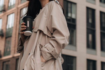 Cropped young woman wearing beige long seasonal trench coat, hand in pocket, walking around the city with a cup of coffee. Street style photo shoot