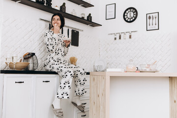 Cheerful woman standing in bright kitchen smiling eat candy, dressed in patterned pajama set. The kitchen counter arranged with ingredients, utensils

