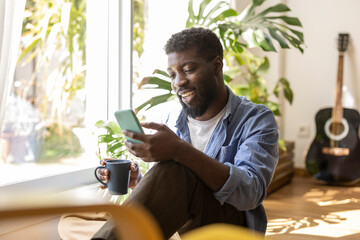 Smiling non-binary person holding coffee cup and using smart phone at home