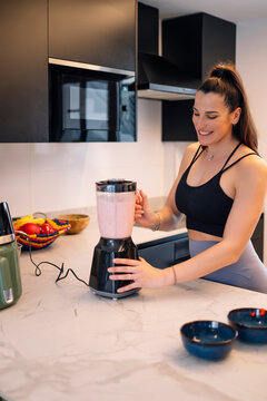 Smiling woman preparing smoothie using blender in kitchen at home