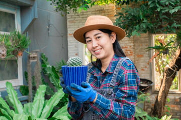 Asian woman gardener standing holding cactus tree smiling with confidence in the backyard. Beautiful woman wears hat gardening at home as hobby, small business owner entrepreneur in shop or backyard.